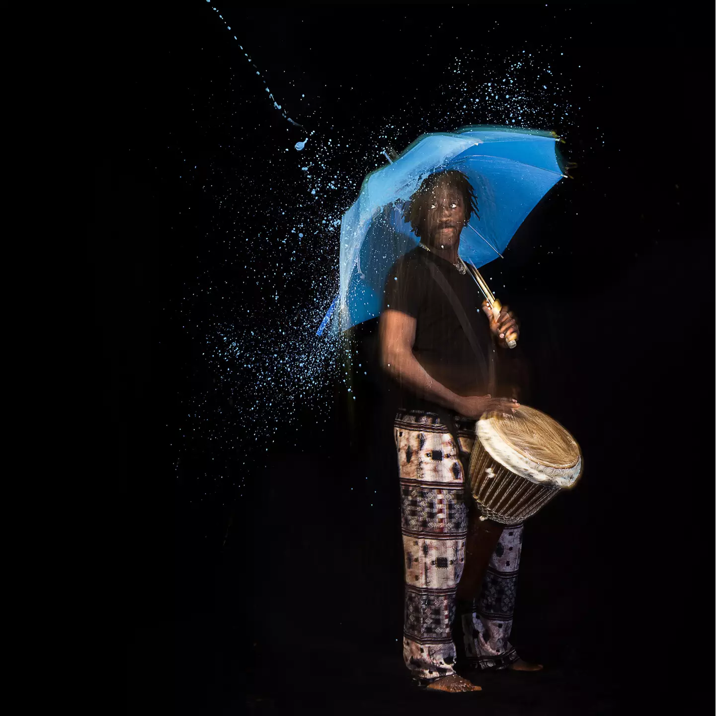Senegalese dancer playing the conga drum while being under the umbrella and hit by stylized blue water from the top.