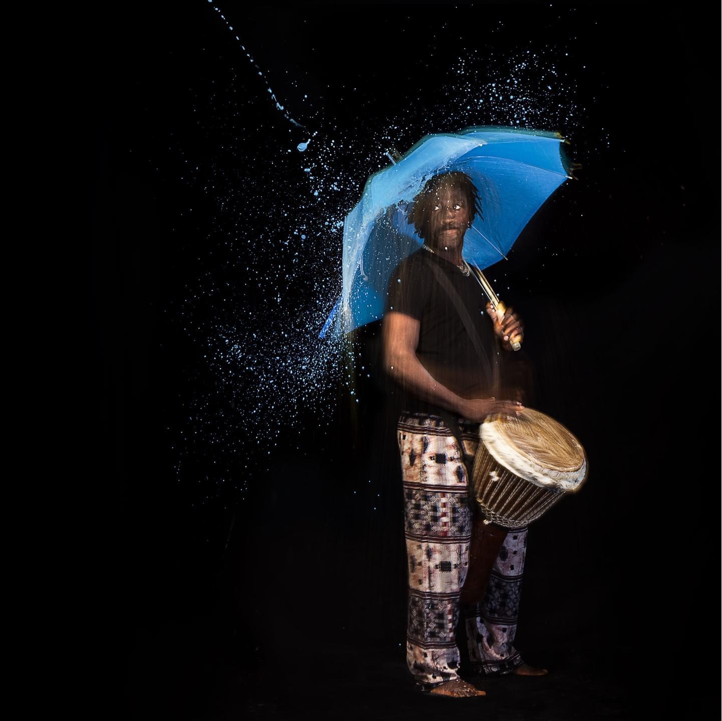 Senegalese dancer playing the conga drum while being under the umbrella and hit by stylized blue water from the top.